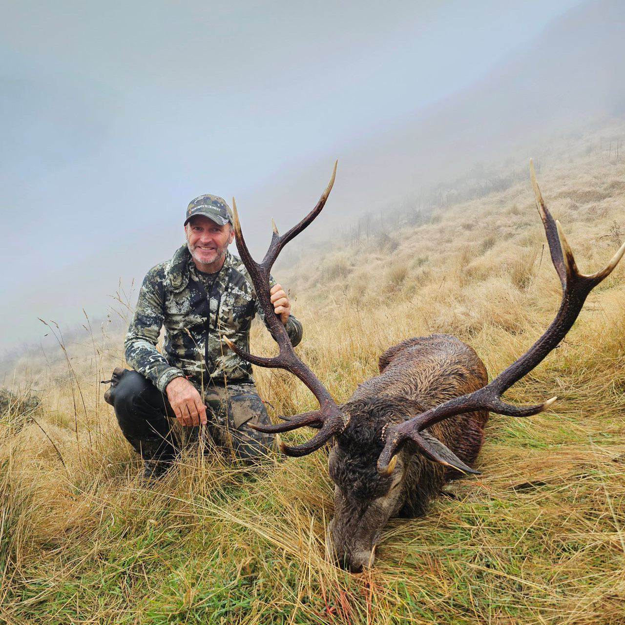 A man sitting beside a stag wearing a zip-up 100% merino hoodie in Tahr Country snow melt camouflage print on the side of a mountain.