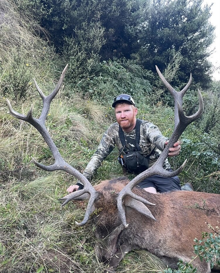 A man sitting beside a stag, wearing a 100% merino long sleeve top in Tahr Country Snow Melt camouflage print.