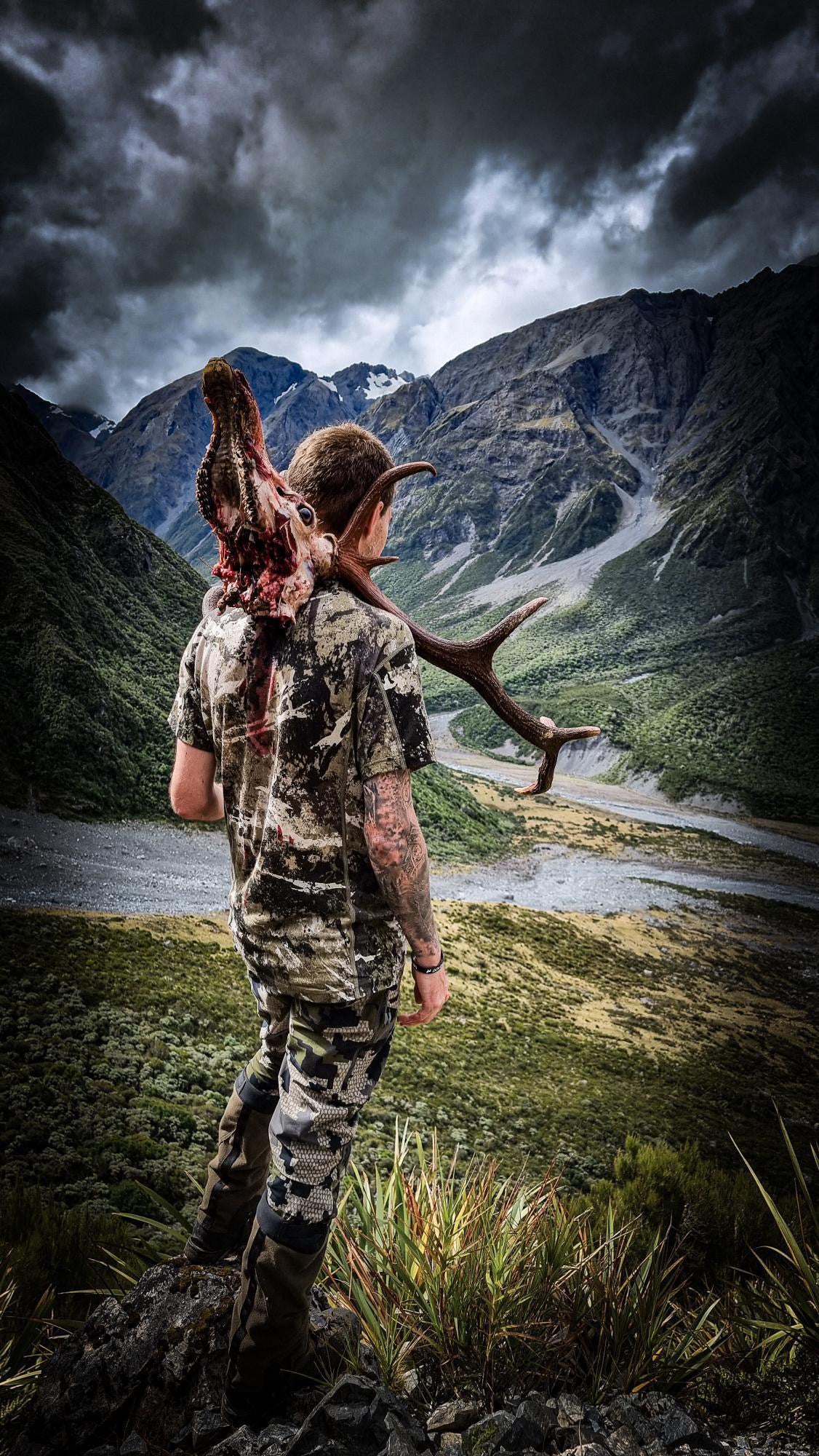 A man carrying a Tahr Head over the back of his shoulder on the side of a mountain wearing a 100% merino short sleeve t-shirt in Tahr Country Snow Melt camouflage print.