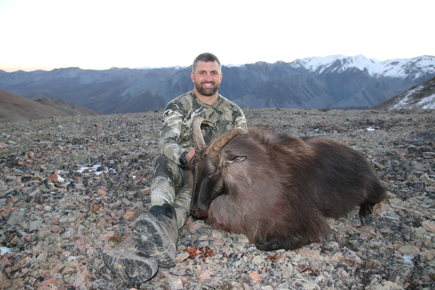 A man sitting beside a Tahr atop a mountain wearing a pair of Tahr Country camouflage patterned long Johns made from a blend of 90% merino wool and 10% polyester, and a tahr country merino long sleeve top.